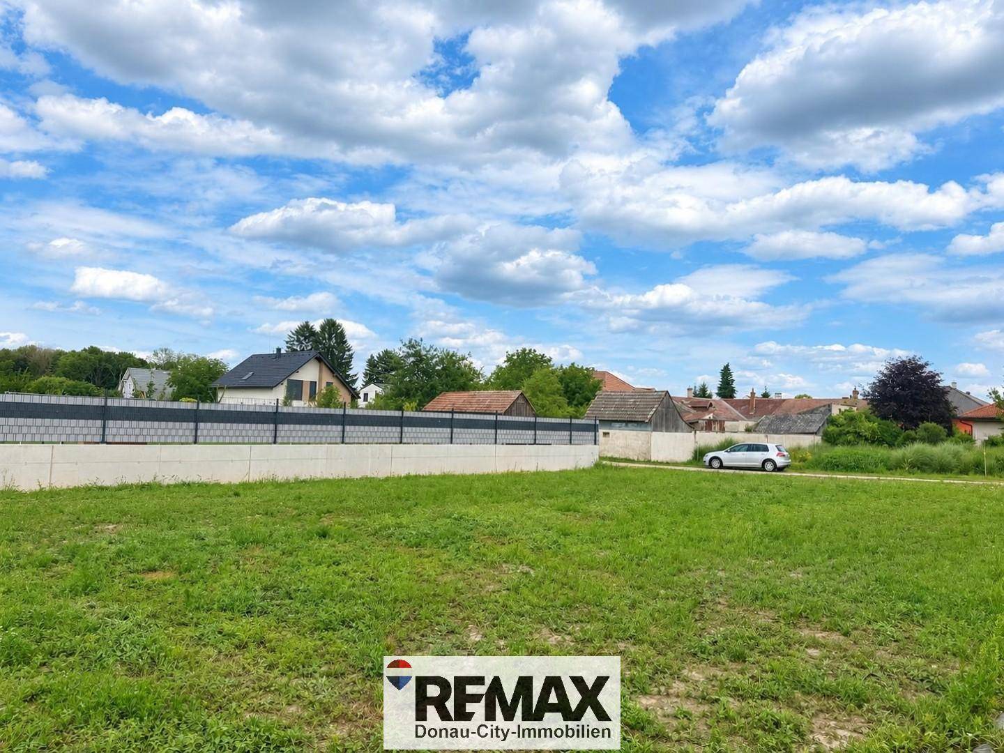 Offenes, sonniges Grundstück mit Grasfläche und Häusern im Hintergrund unter einem bewölkten Himmel.