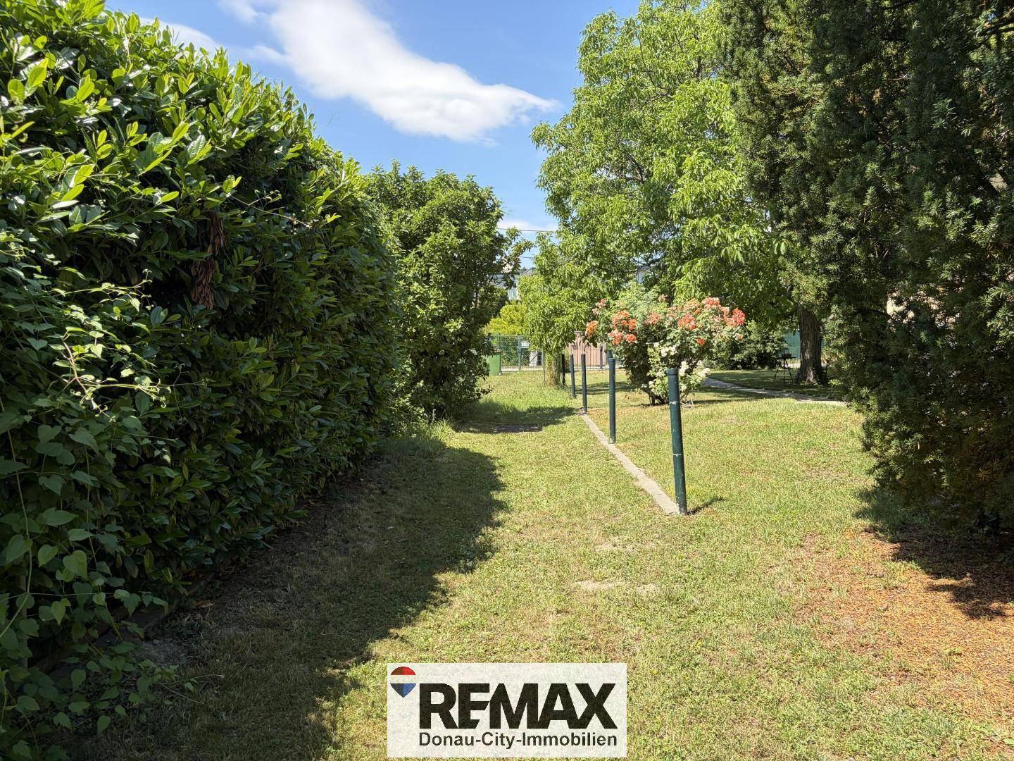 Ein schmaler Gartenweg, gesäumt von hohen Hecken und Bäumen, führt durch eine grüne Landschaft.