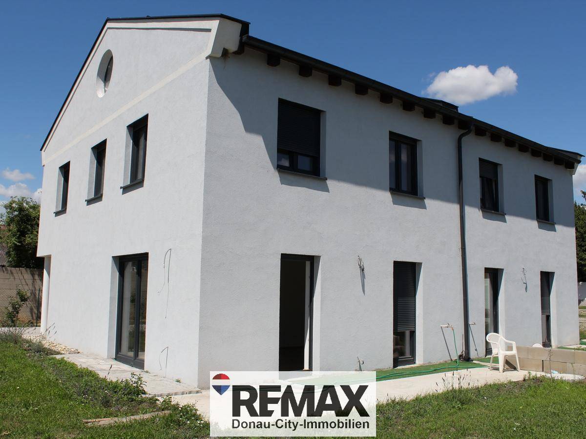 Moderne Hausfassade in Weiß mit dunklen Fensterrahmen und Rollläden, umgeben von Grünflächen.