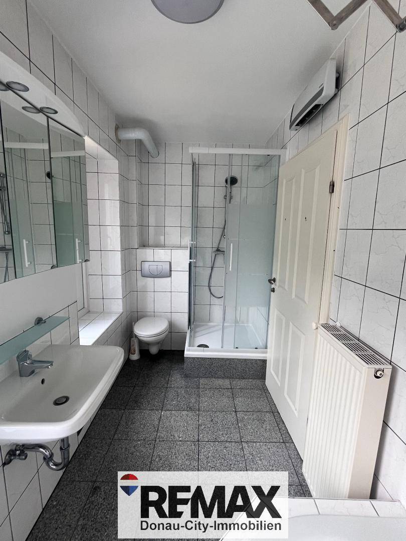 Badezimmer mit weißen Wandfliesen, grauen Bodenfliesen, Dusche und WC.