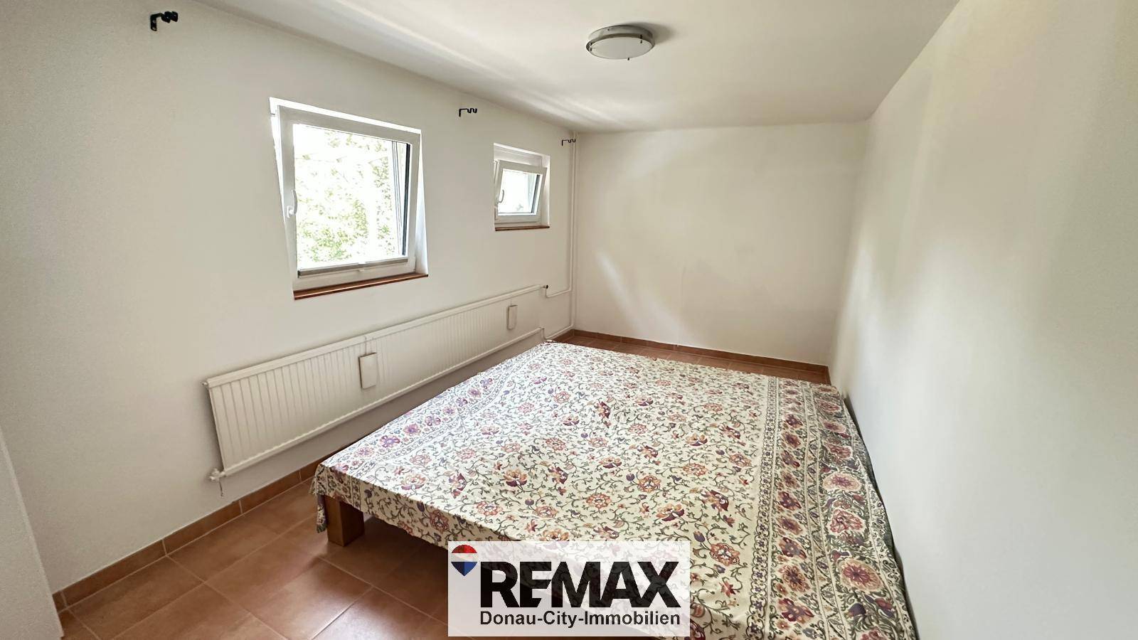 Schlafzimmer mit Fliesenboden, zwei Fenstern und einem großen Bett.
