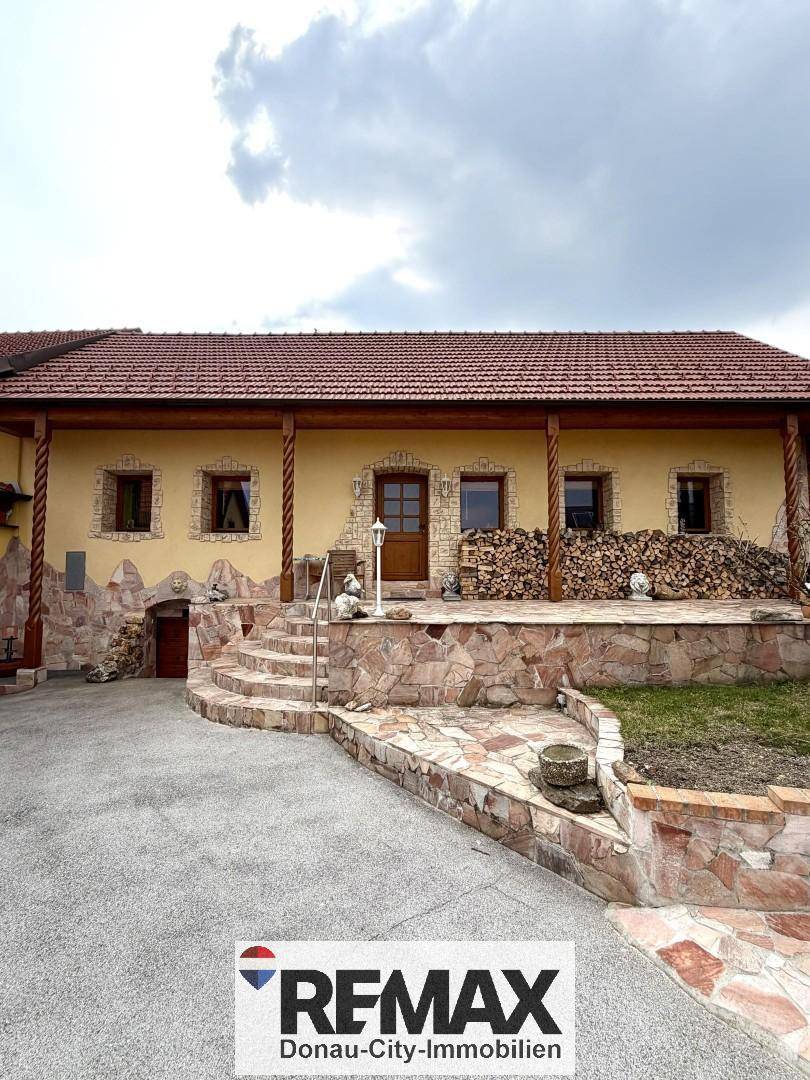 Vorderansicht des Hauses mit gelber Fassade, Steindetails, überdachtem Eingangsbereich und Holzstapel.