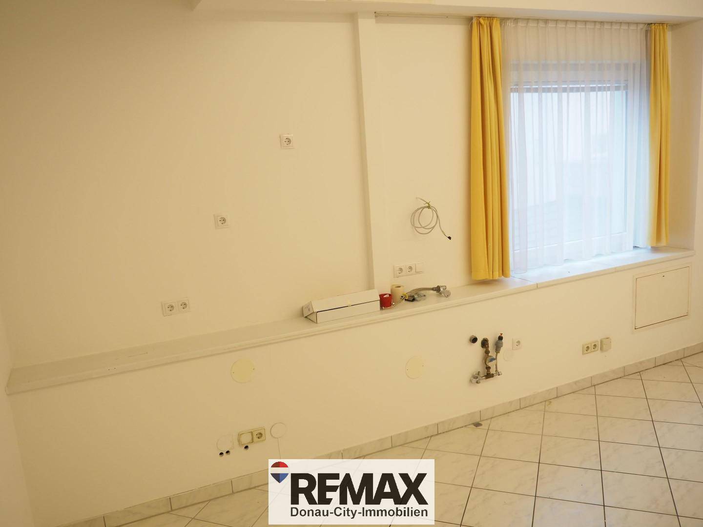 Büroraum mit Fliesenboden, einem Fenster und sichtbaren Installationen an der Wand, die auf Umbauarbeiten hindeuten.