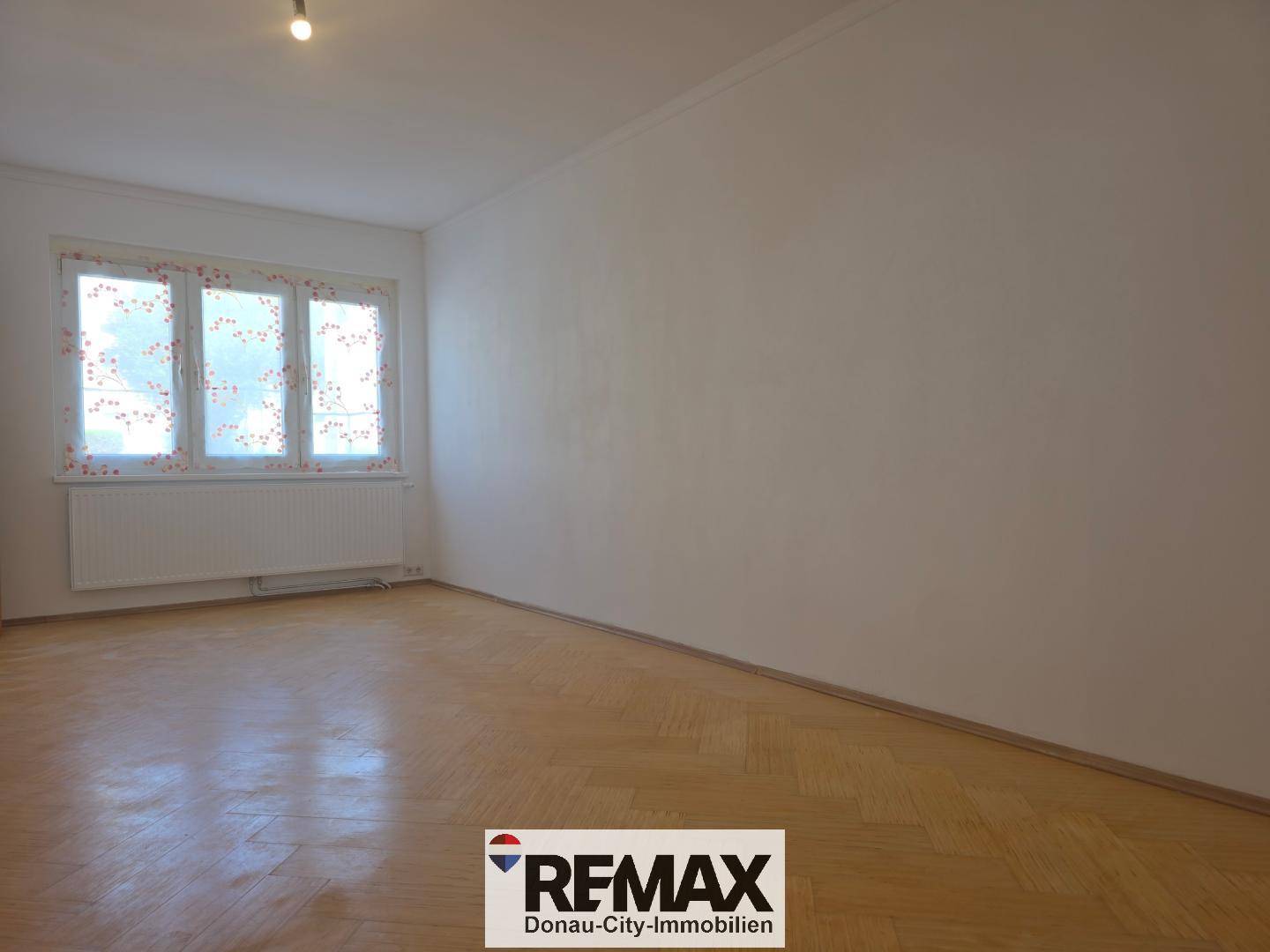 Leeres Wohnzimmer mit Fischgrätparkett und großen Fenstern, die viel Tageslicht hereinlassen.