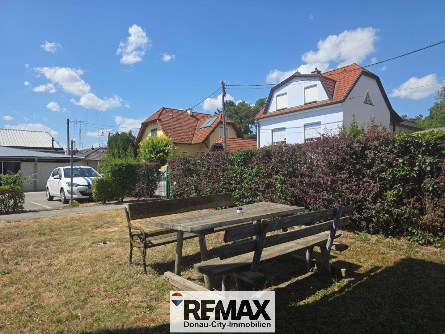 Gemütlicher Gartenbereich mit Holztisch und Bänken, umgeben von Hecken und Bäumen unter blauem Himmel.