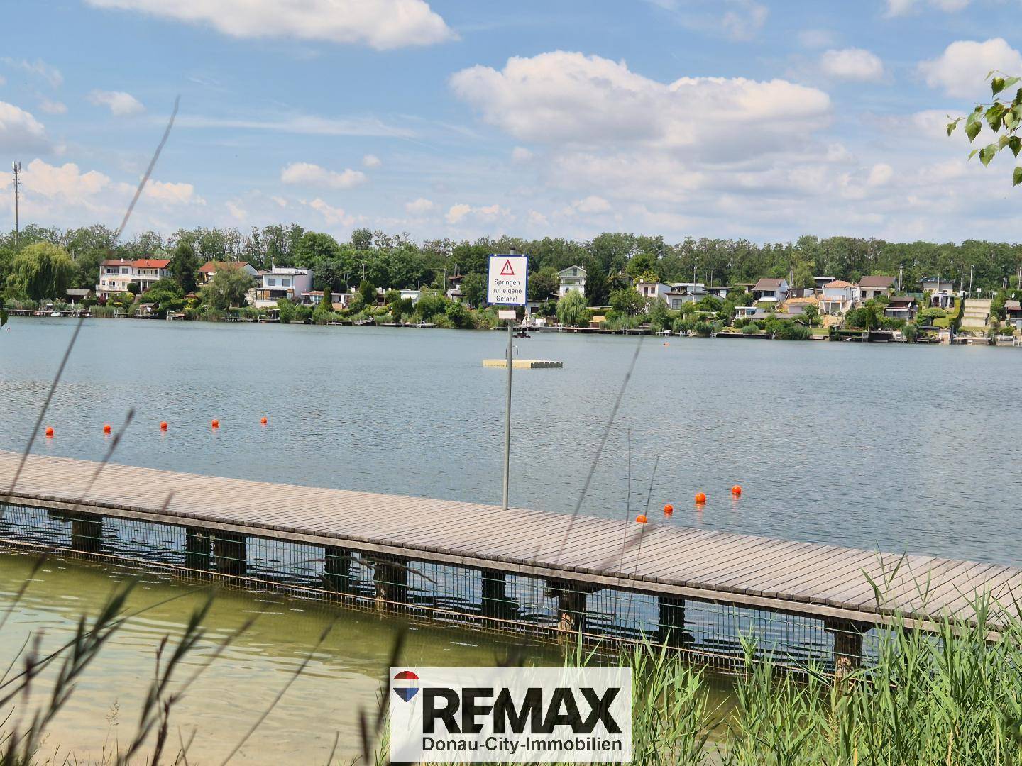 Idyllischer Blick über einen See mit Holzanlegesteg und Häusern am gegenüberliegenden Ufer unter blauem Himmel.