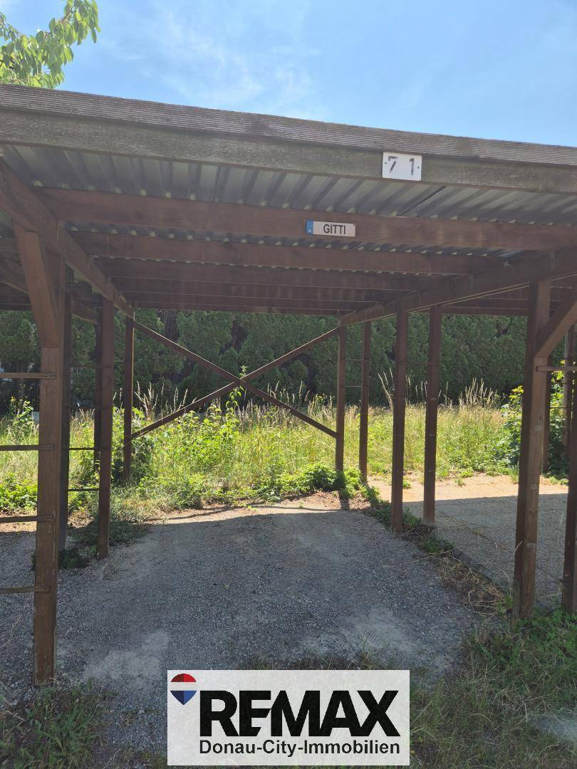 Einfacher Holzcarport mit Wellblechdach und Schotterboden, bietet Schutz für ein Fahrzeug.