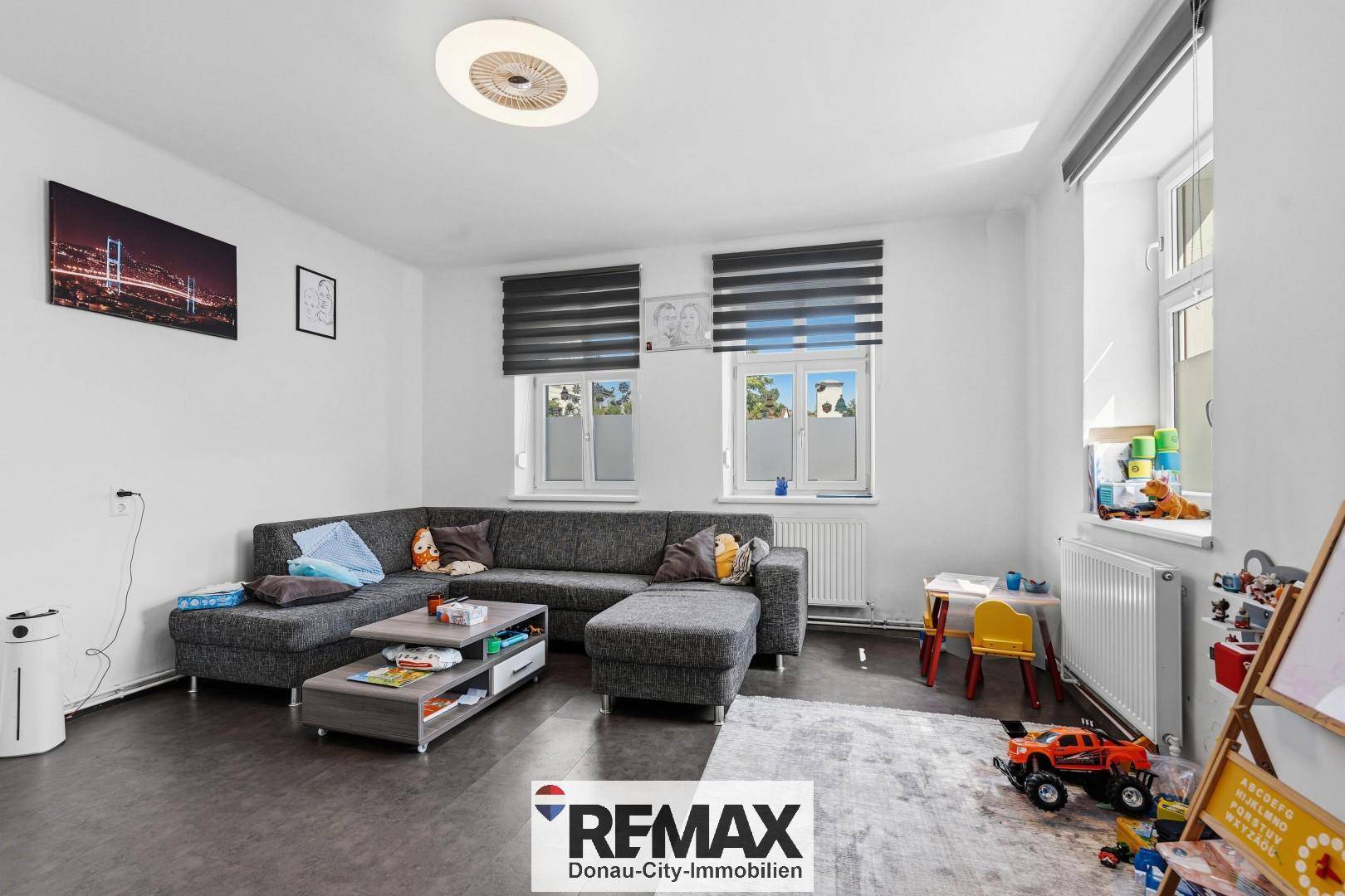 Helles Wohnzimmer mit großem Ecksofa, Spielbereich für Kinder und Fenstern mit Jalousien.