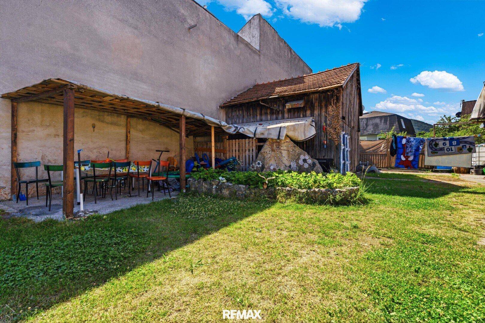 Gartenbereich mit überdachtem Sitzplatz, Rasen und Nebengebäuden.