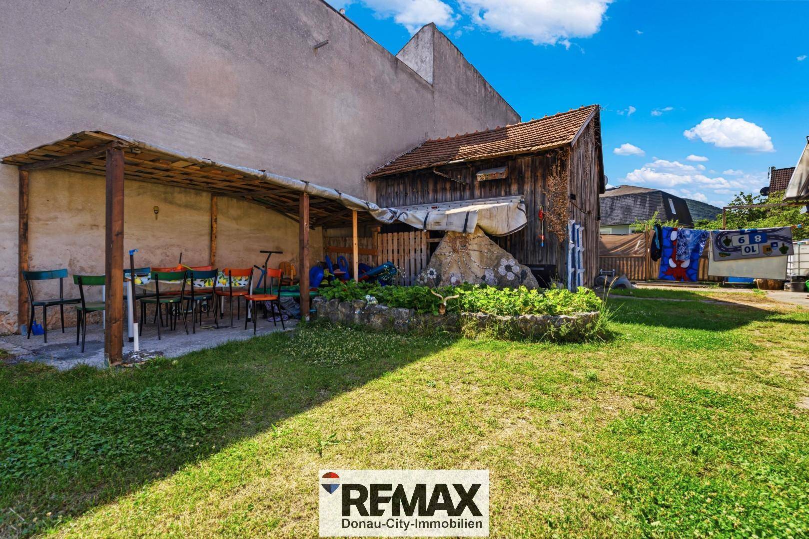 Gartenbereich mit Rasen, überdachtem Sitzplatz und einem alten Holzschuppen im Hintergrund.