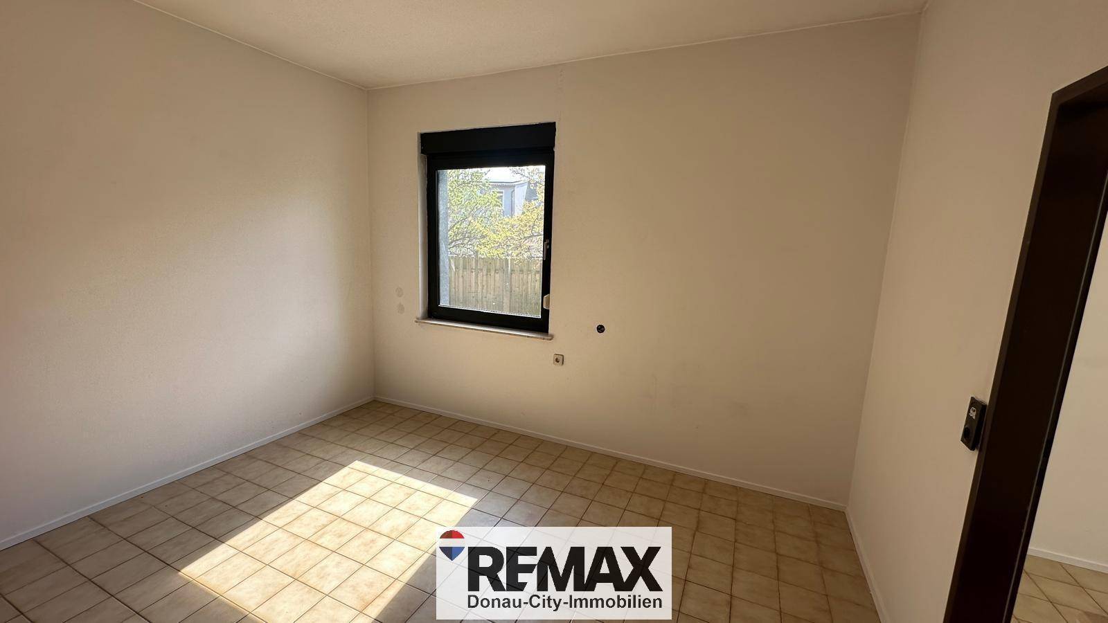 Helles Schlafzimmer mit Fliesenboden und Fenster mit Blick ins Grüne.