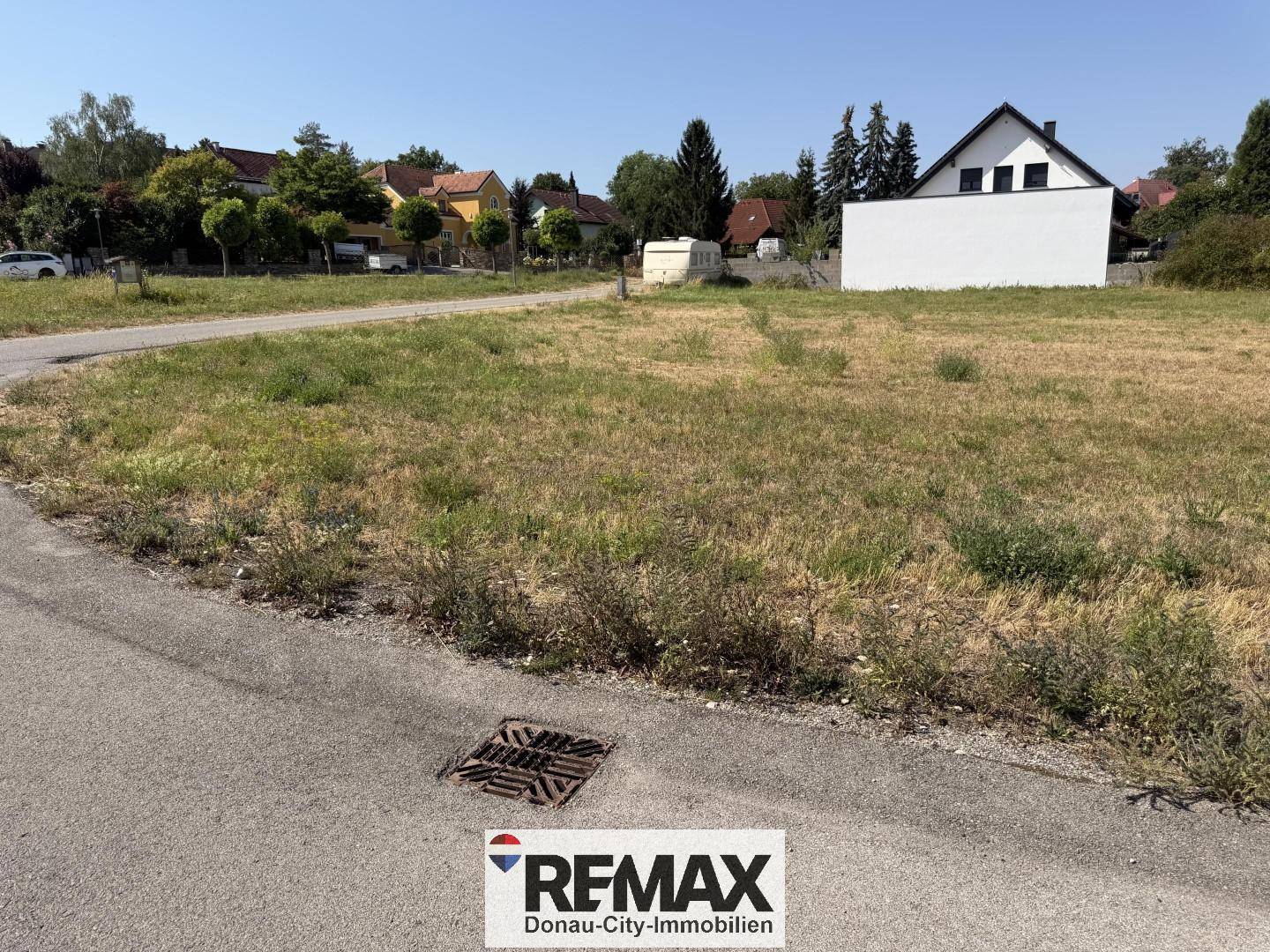 Unbebautes Grundstück mit angrenzender Straße und Wohngebäuden im Hintergrund unter blauem Himmel.