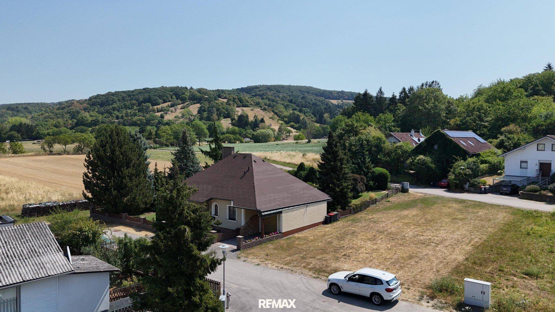 Luftaufnahme eines Einfamilienhauses mit großem Garten und bewaldeten Hügeln im Hintergrund.