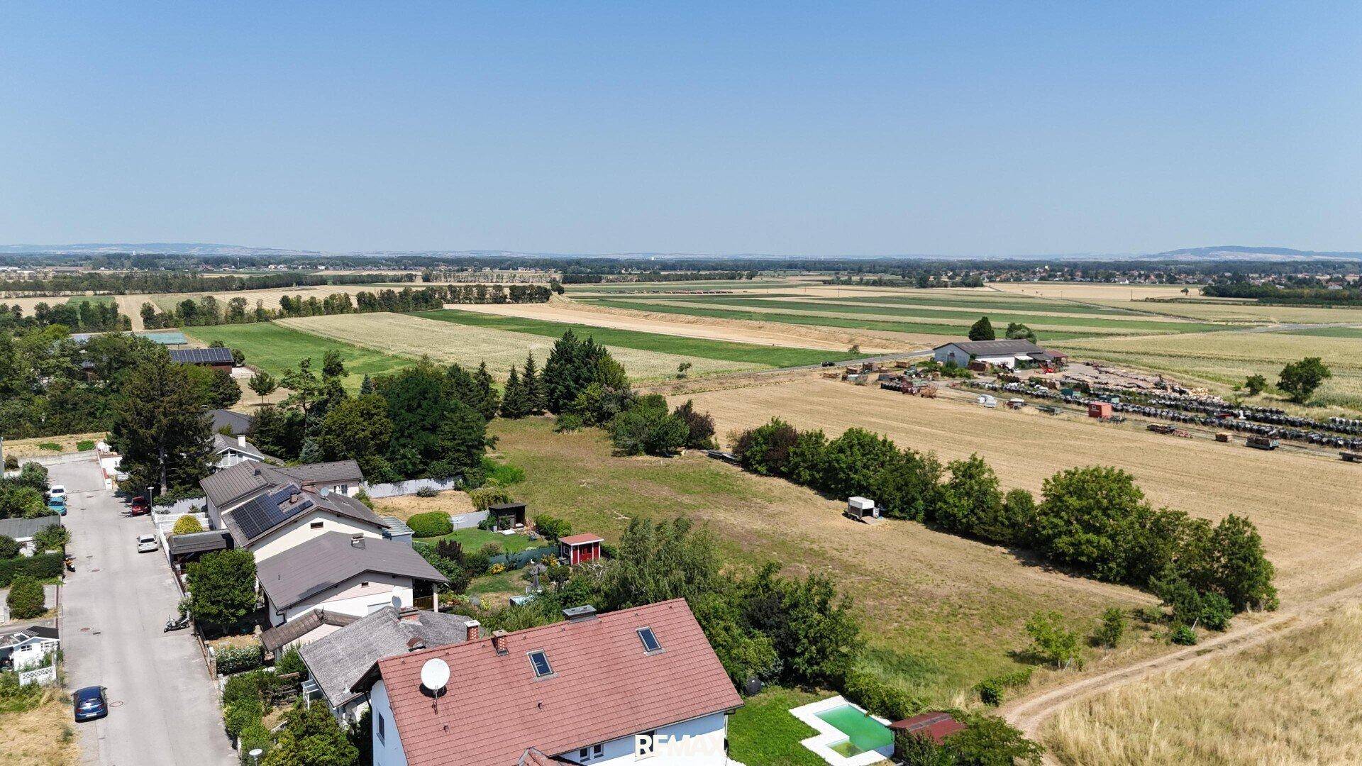 Panoramablick über eine Wohnsiedlung und weite landwirtschaftliche Flächen unter strahlendem Sonnenschein.
