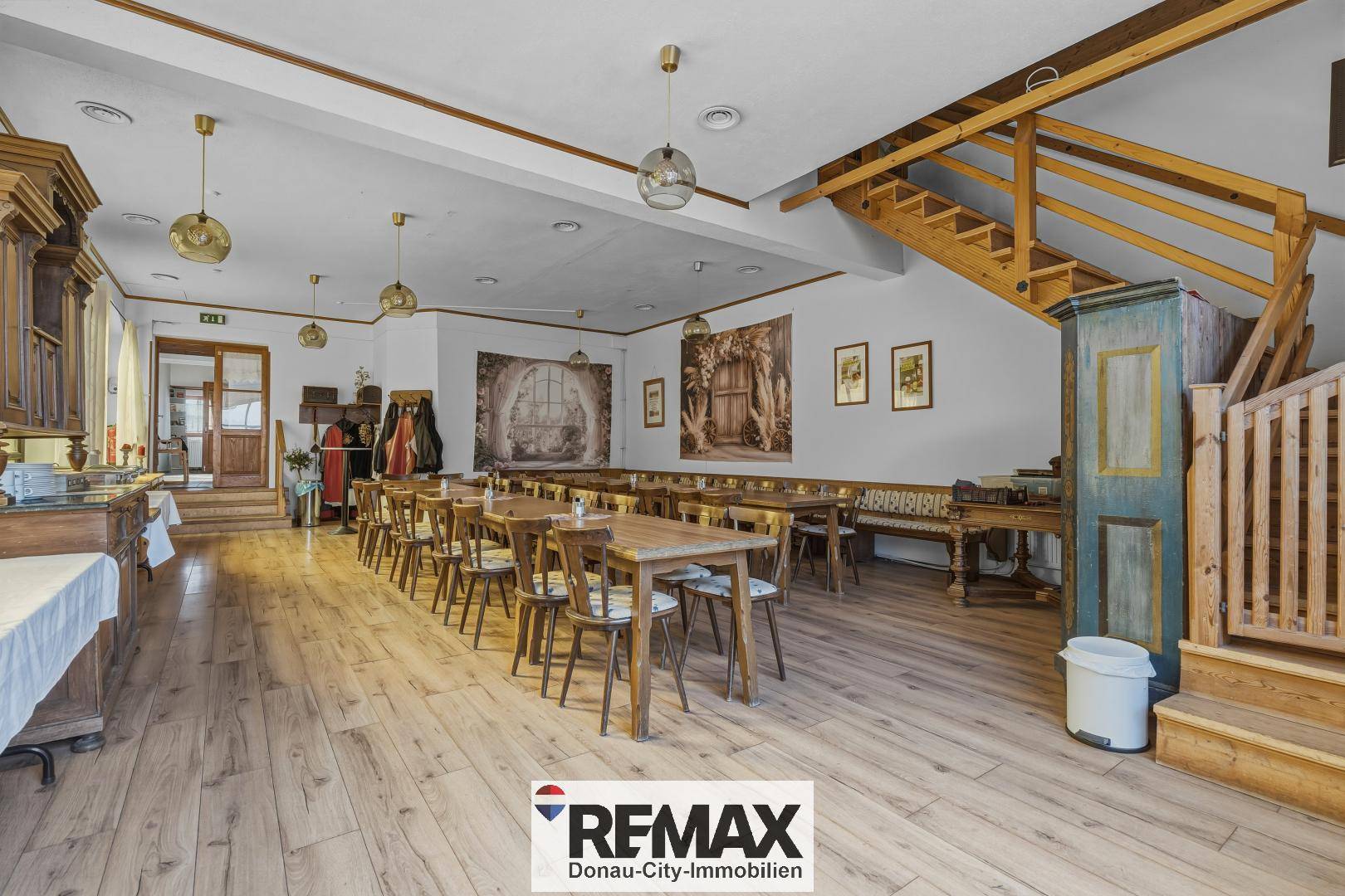 Geräumiger Saal mit Holzboden, langen Tischen und einer Holztreppe zum Obergeschoss.