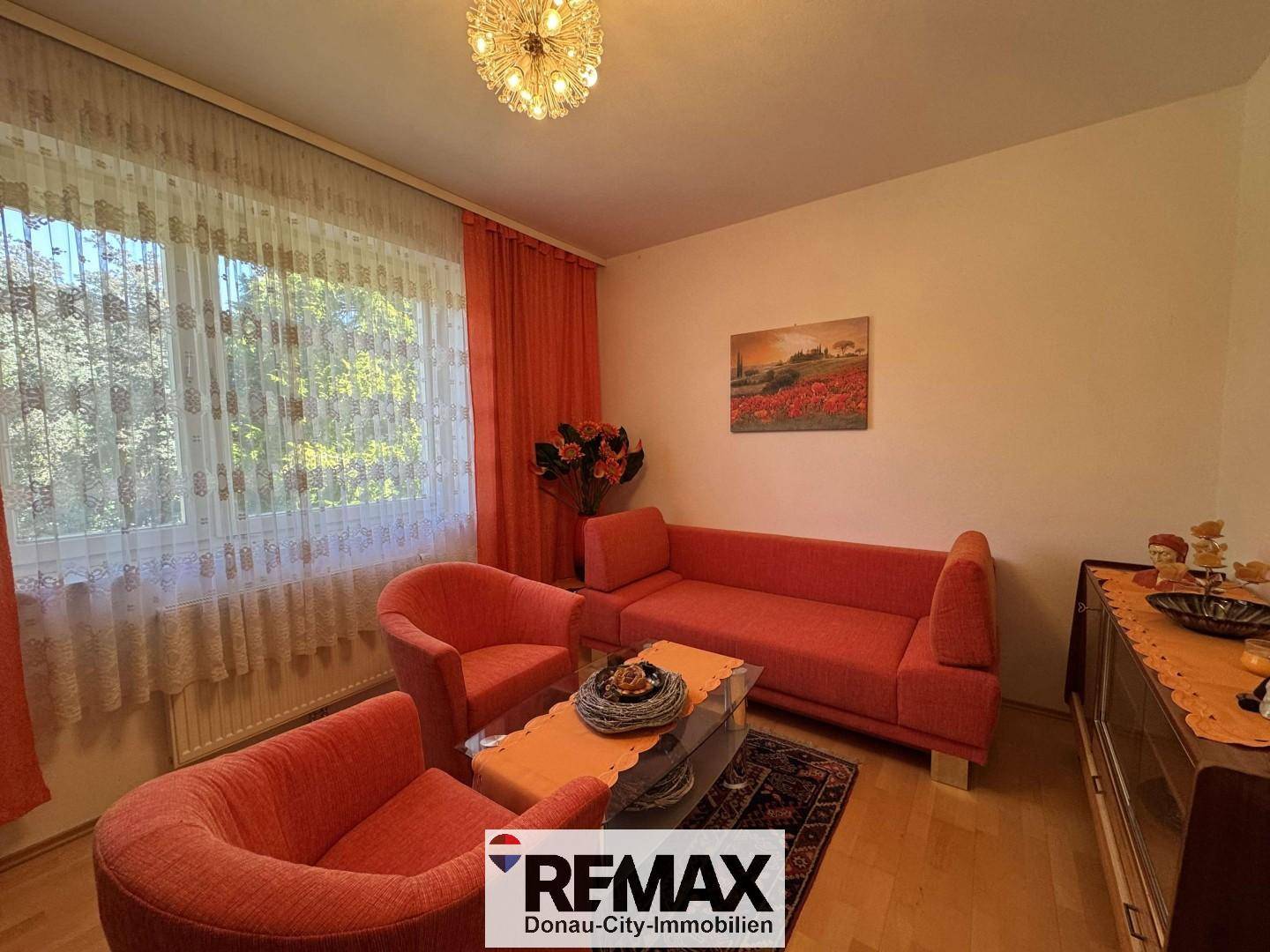 Helles Wohnzimmer mit orangefarbenen Sofas, Holzboden und großen Fenstern mit Vorhängen.