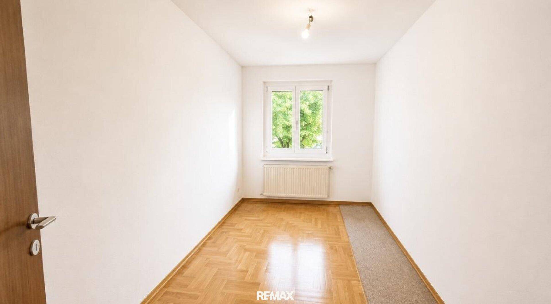 Helles Zimmer mit Fischgrätparkett, weißen Wänden und einem Fenster mit Blick ins Grüne.