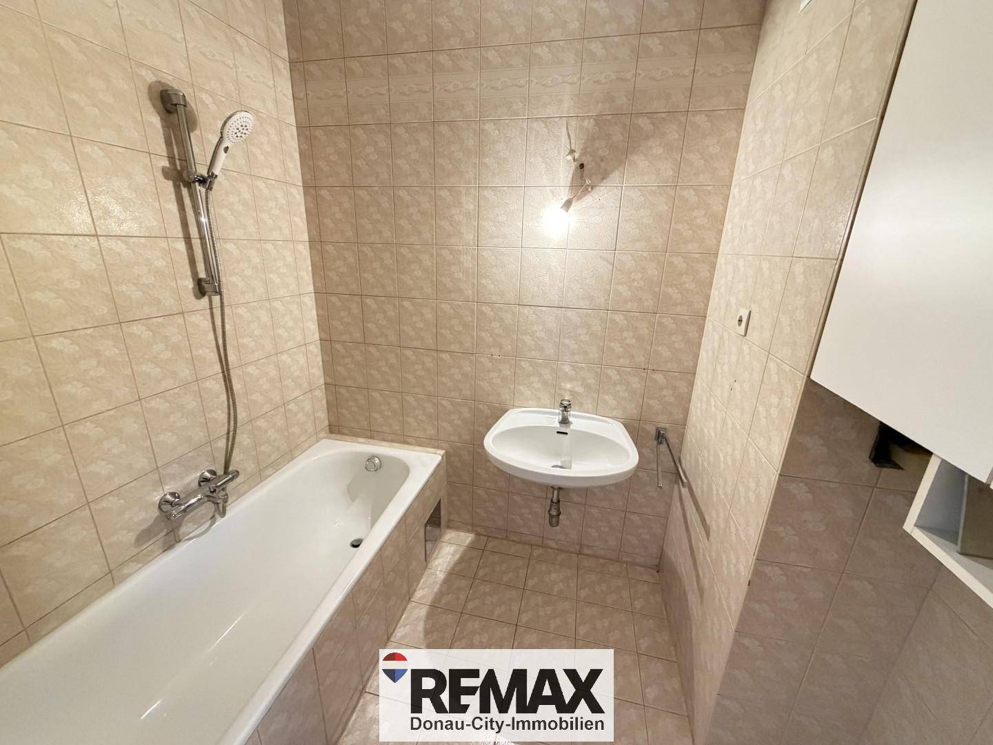 Badezimmer mit beigefarbenen Fliesen, einer Badewanne mit Duschfunktion und einem einzelnen Waschbecken.