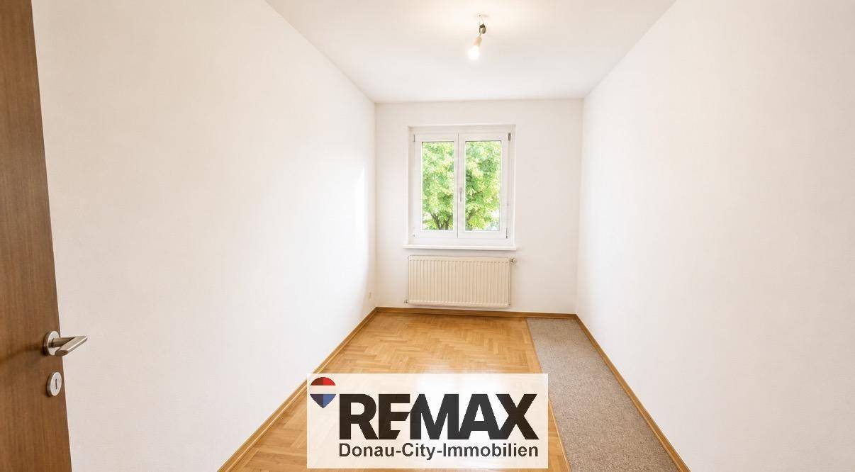 Helles Zimmer mit Parkettboden und Teppichbereich, weißen Wänden und einem Fenster mit Grünblick.