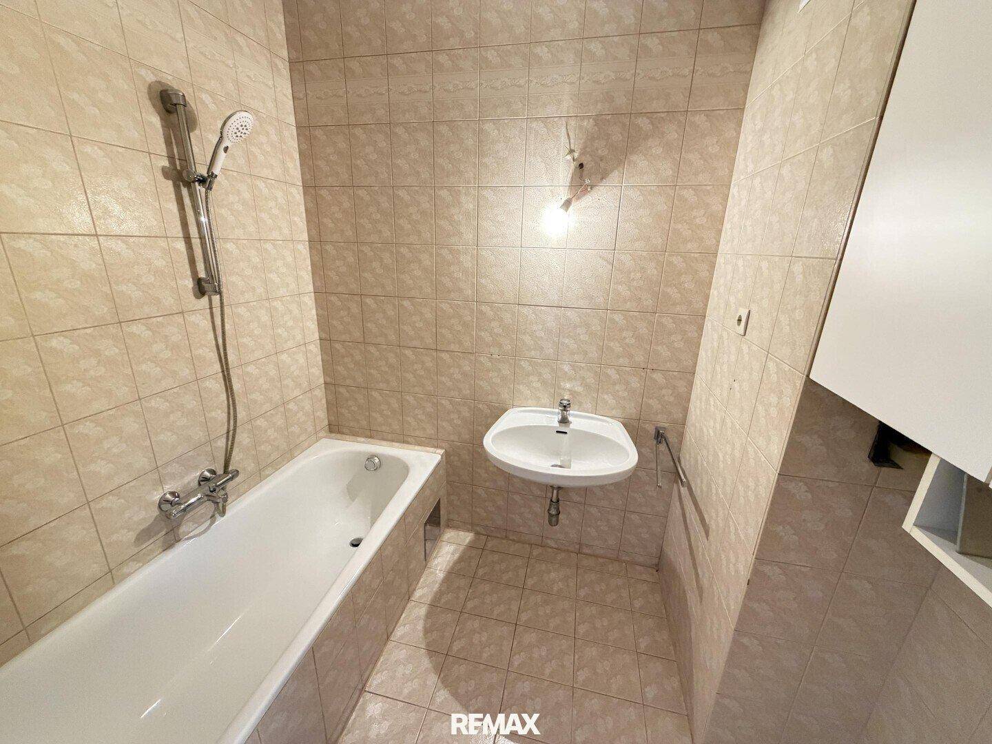 Badezimmer mit beigefarbenen Fliesen, Badewanne mit Duschfunktion und einem einzelnen Waschbecken.