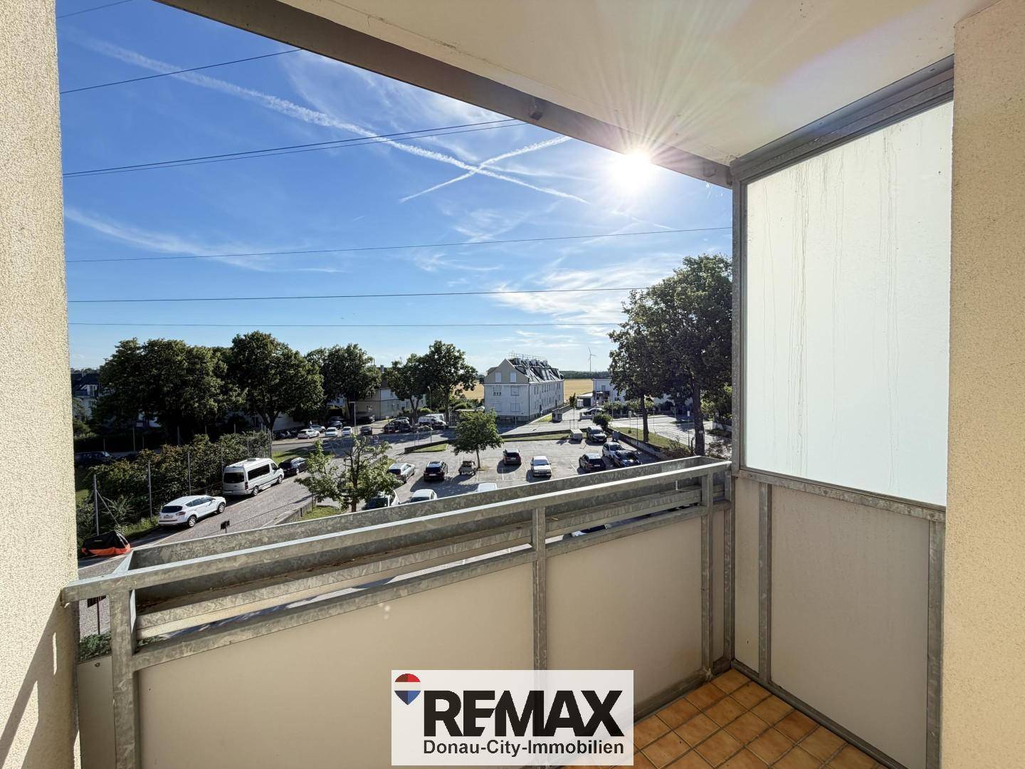 Sonniger Balkon mit Fliesenboden und Blick auf die Straße sowie umliegende Bäume.
