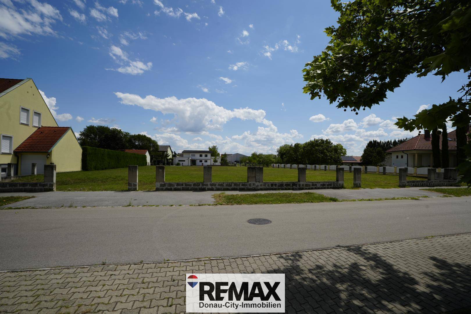 Blick von der Straße auf ein grünes Grundstück mit niedriger Betonmauer und umliegenden Häusern.
