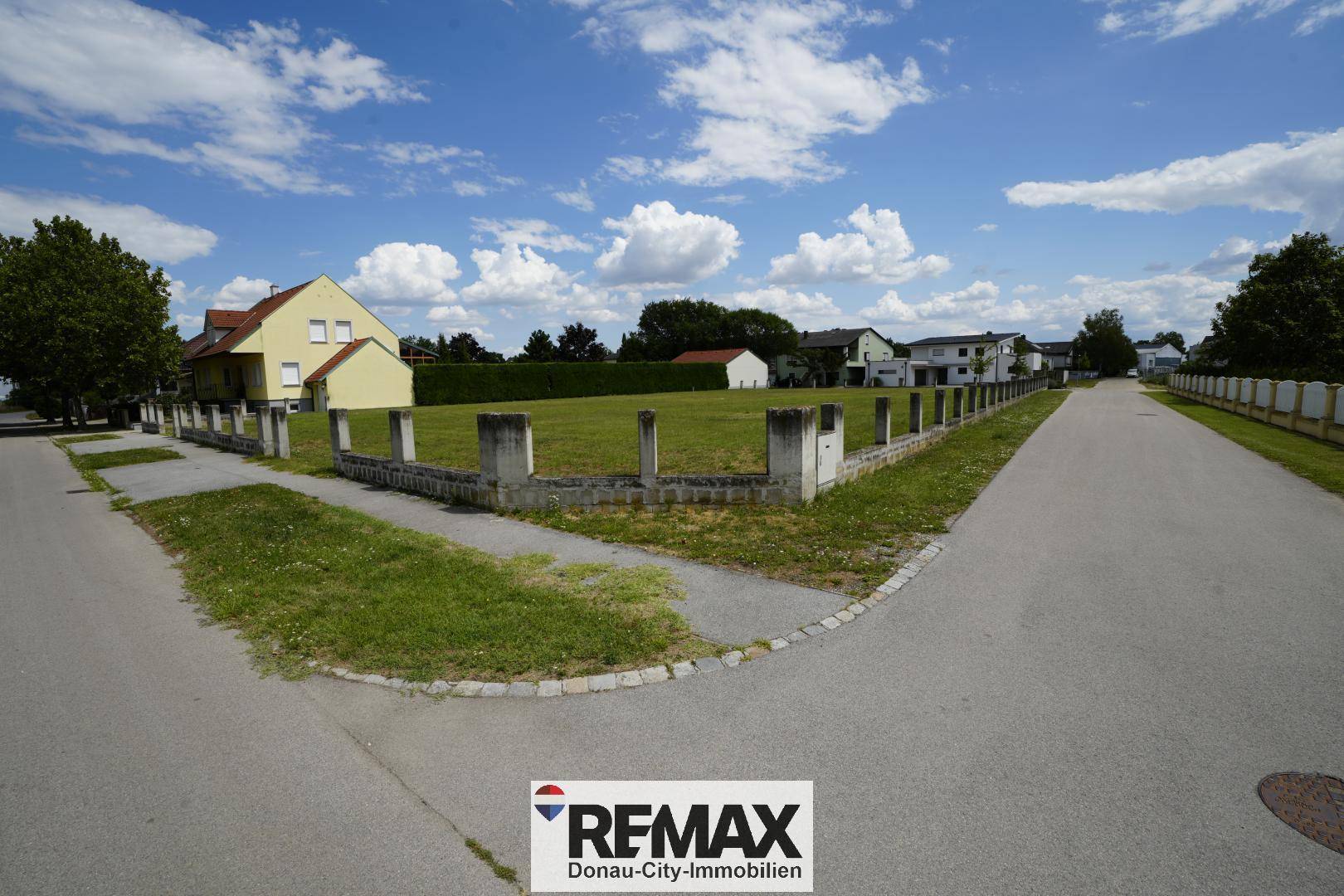 Eckansicht des grünen Grundstücks von der Straße aus mit niedriger Begrenzung und umliegenden Häusern.