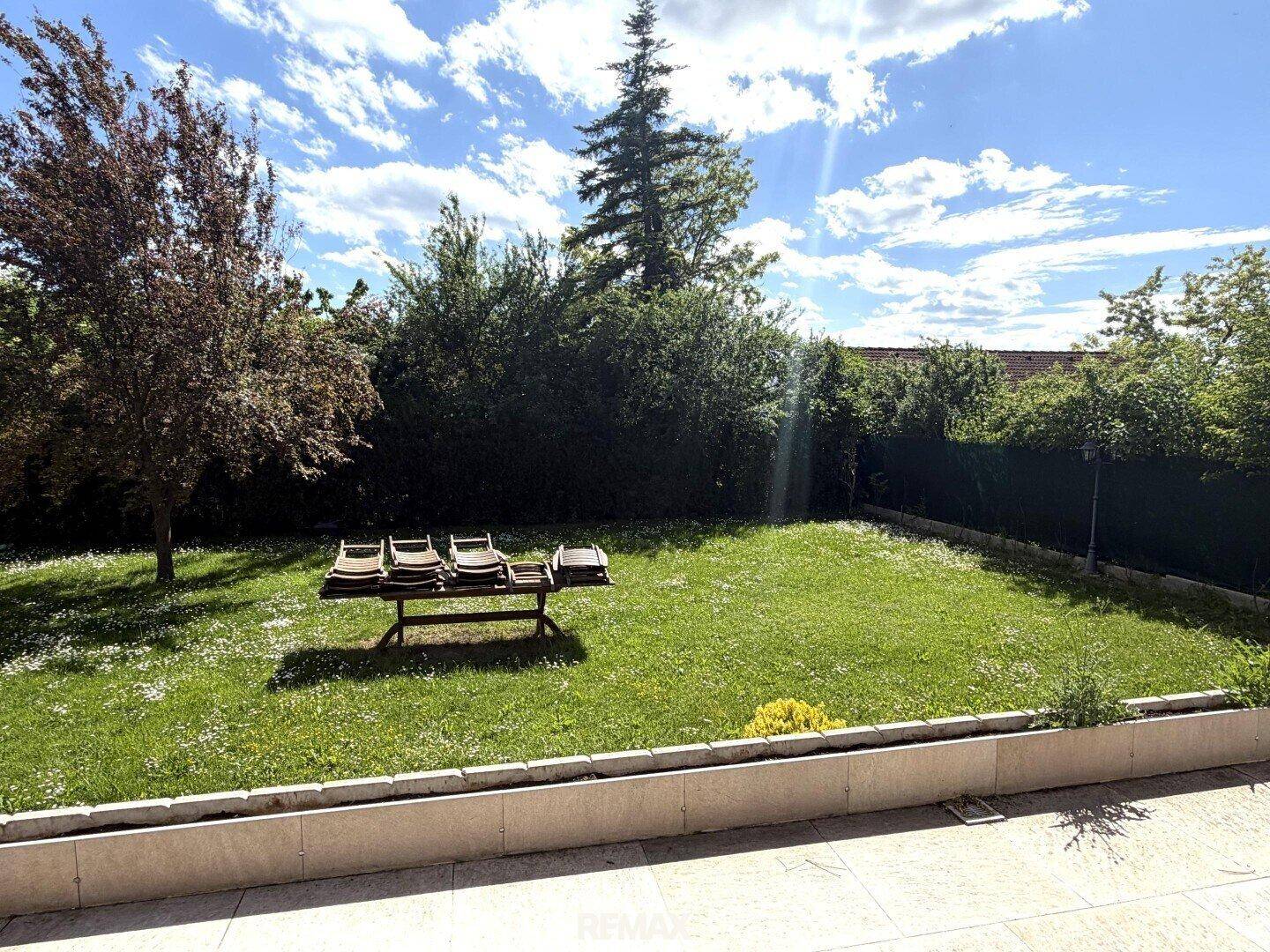 Gepflegter Garten mit Rasenfläche, Bäumen und Liegestühlen unter blauem Himmel.