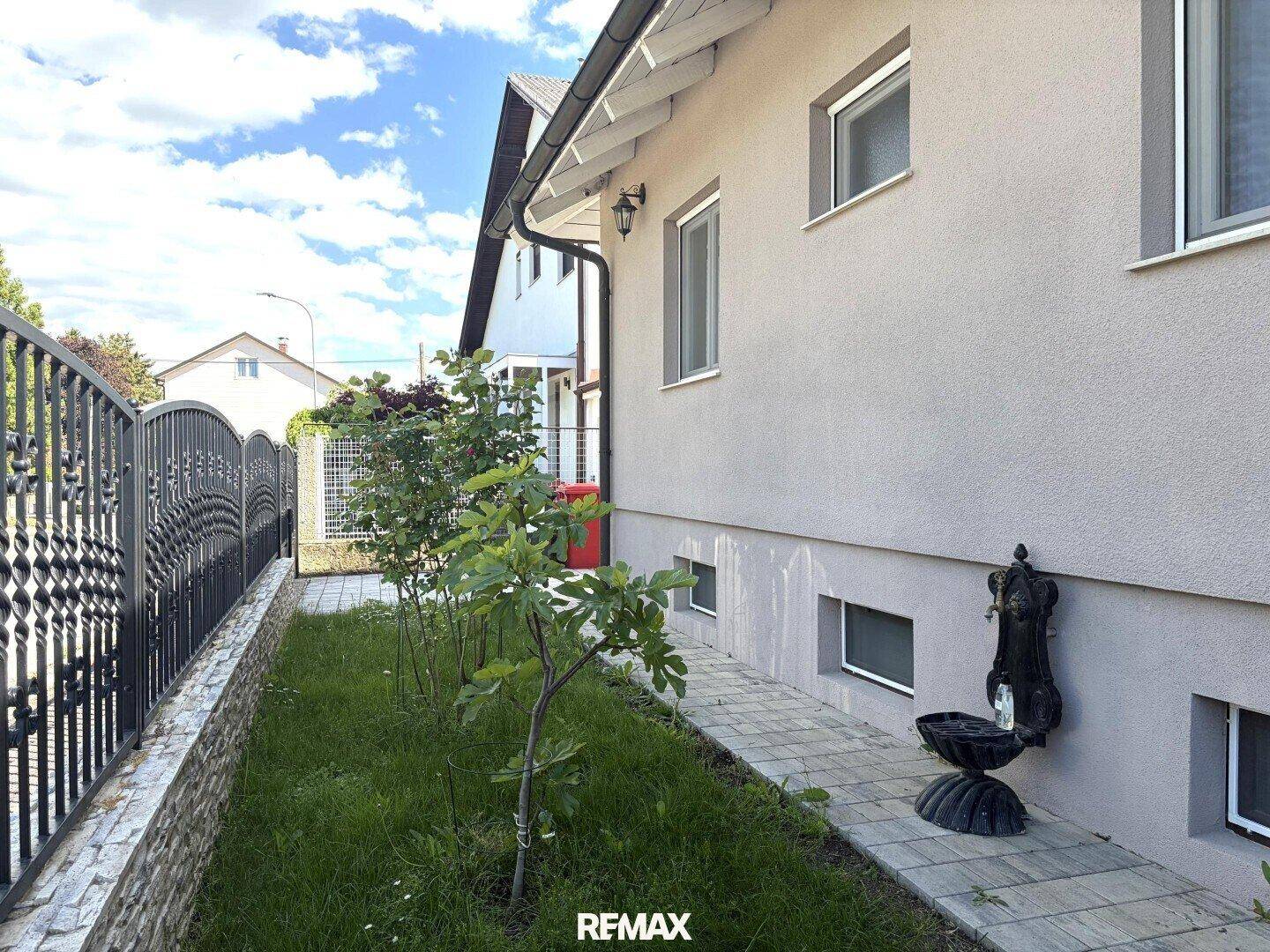 Seitenansicht des Hauses mit gepflastertem Weg, kleinem Garten und dekorativem Brunnen.