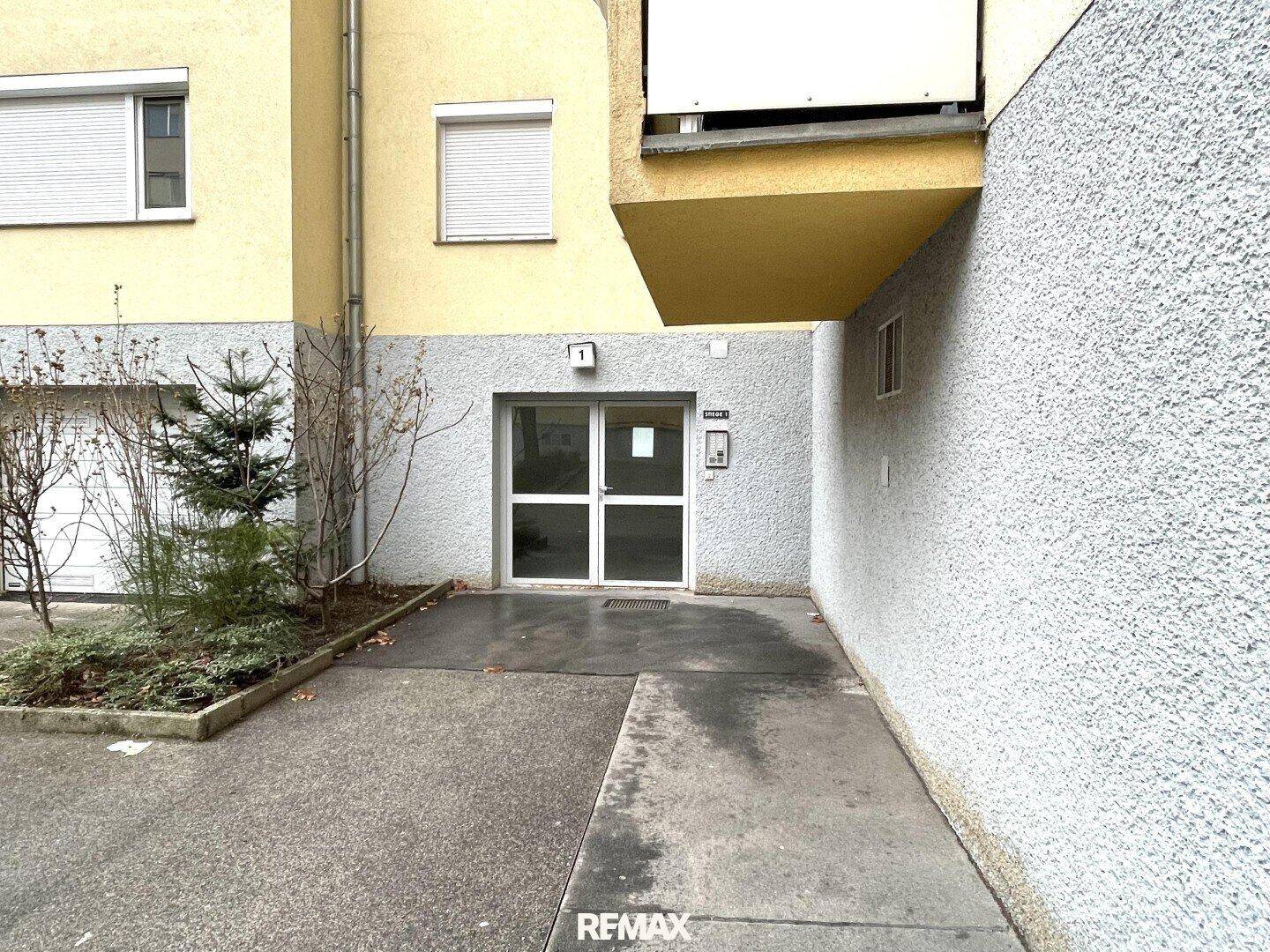 Eingangsbereich eines Wohngebäudes mit moderner Fassade und kleinen Bäumen, einladend gestaltet.