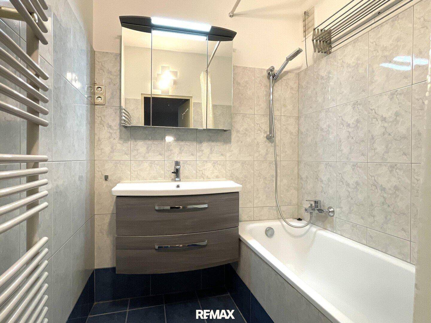 Badezimmer mit Badewanne, Waschbeckenunterschrank und hellen Wandfliesen, inklusive Handtuchheizkörper.