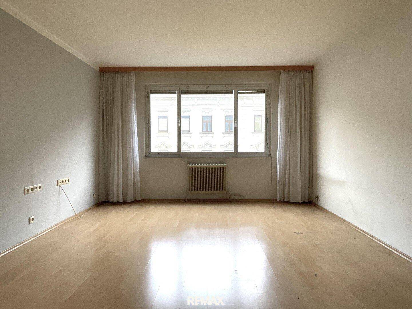 Geräumiges Zimmer mit hellem Parkettboden, großem Fenster und Vorhängen, bietet viel Tageslicht.