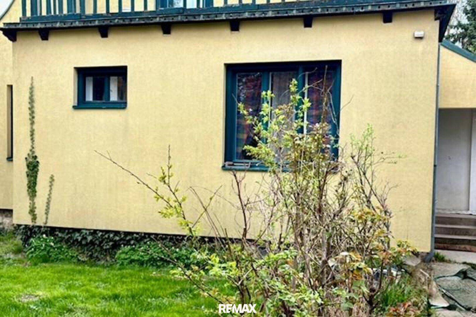 Seitenansicht des Hauses mit gelber Fassade und dunklen Fensterrahmen, umgeben von Grünflächen.