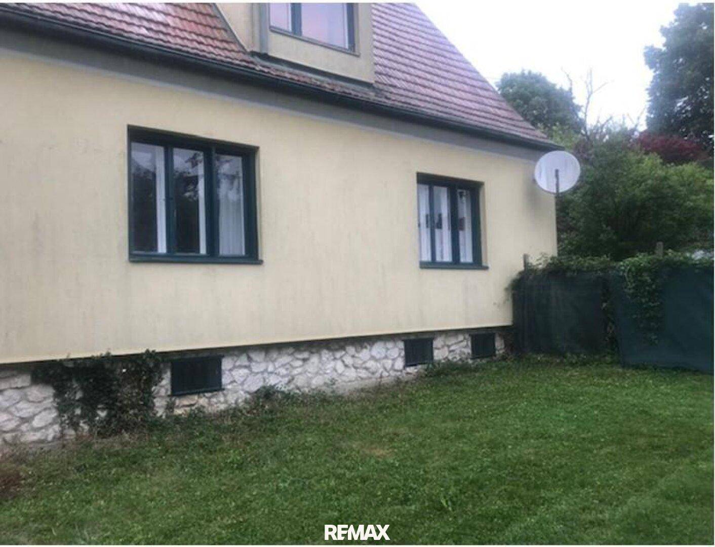 Rückseite des Hauses mit gelber Fassade, Ziegeldach und einem Steinsockel, umgeben von Rasen.