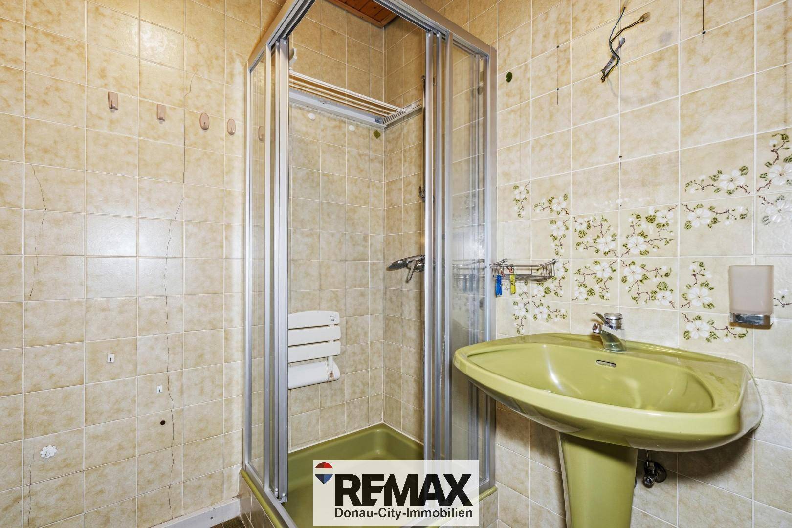 Badezimmer mit Duschkabine, grünem Waschbecken und beigefarbenen Fliesen im 80er-Jahre-Stil.