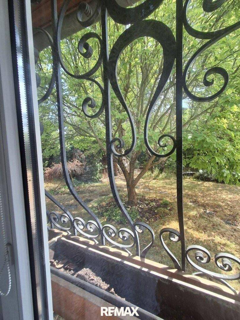 Blick aus dem Fenster mit schmiedeeisernem Gitter auf grüne Bäume und Garten.