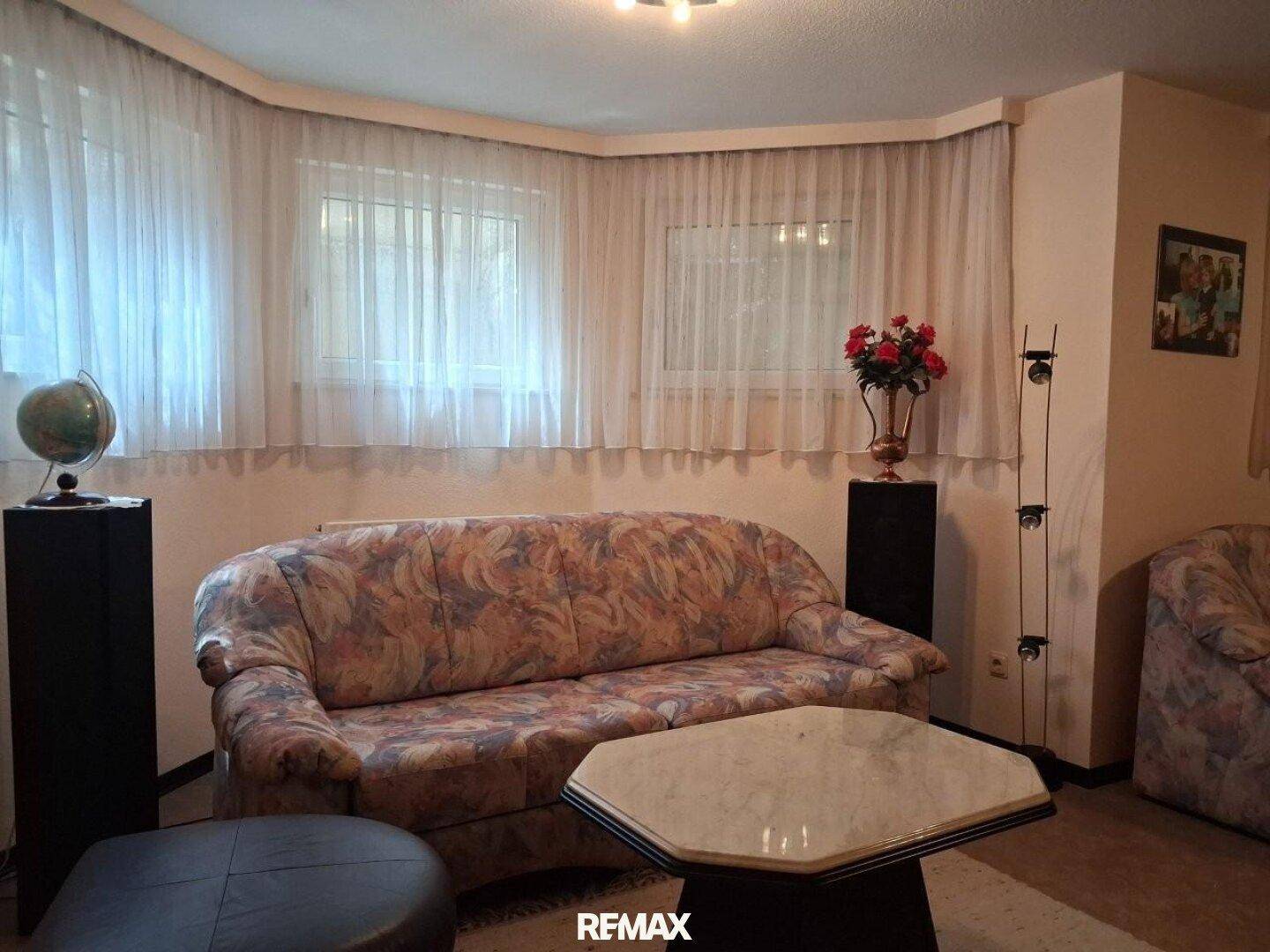 Wohnzimmer mit gemustertem Sofa, Couchtisch und mehreren Fenstern mit Vorhängen.