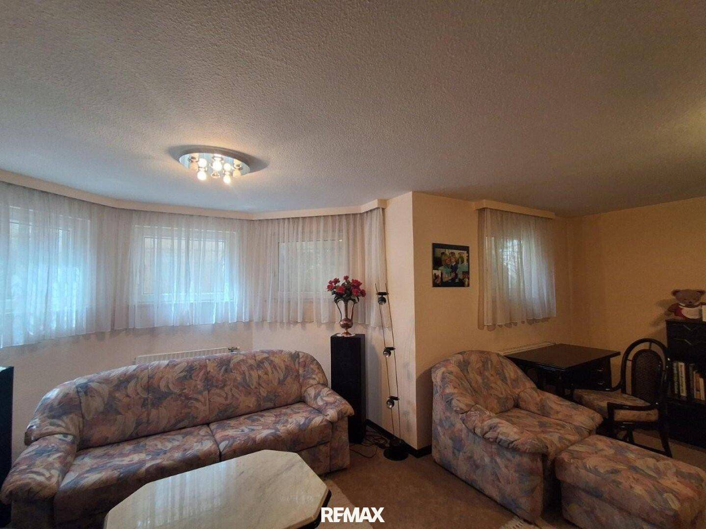 Gemütliches Wohnzimmer mit älteren Möbeln, Teppichboden und mehreren Fenstern.