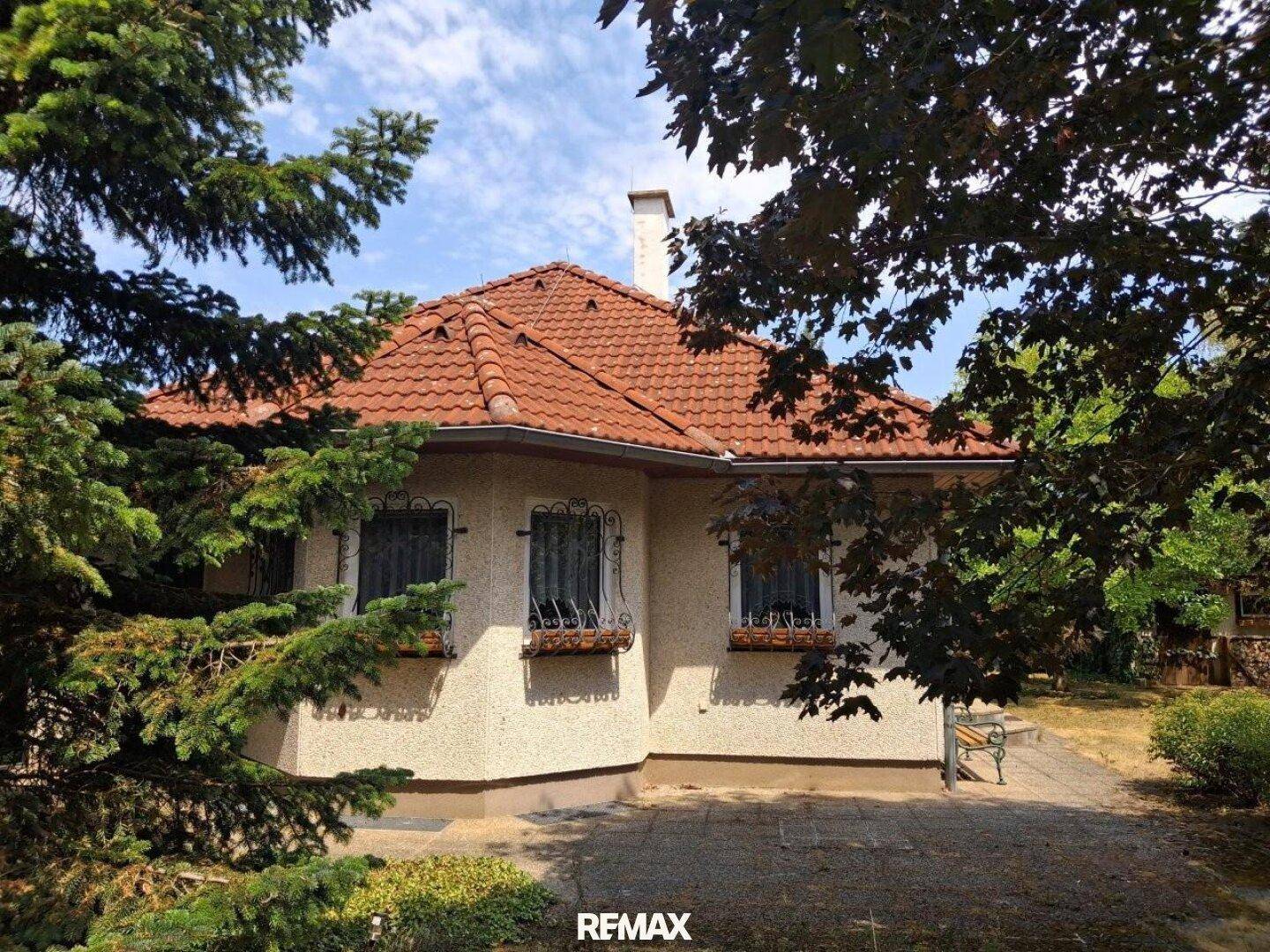 Einfamilienhaus mit rotem Ziegeldach, umgeben von Bäumen und Sträuchern an einem sonnigen Tag.