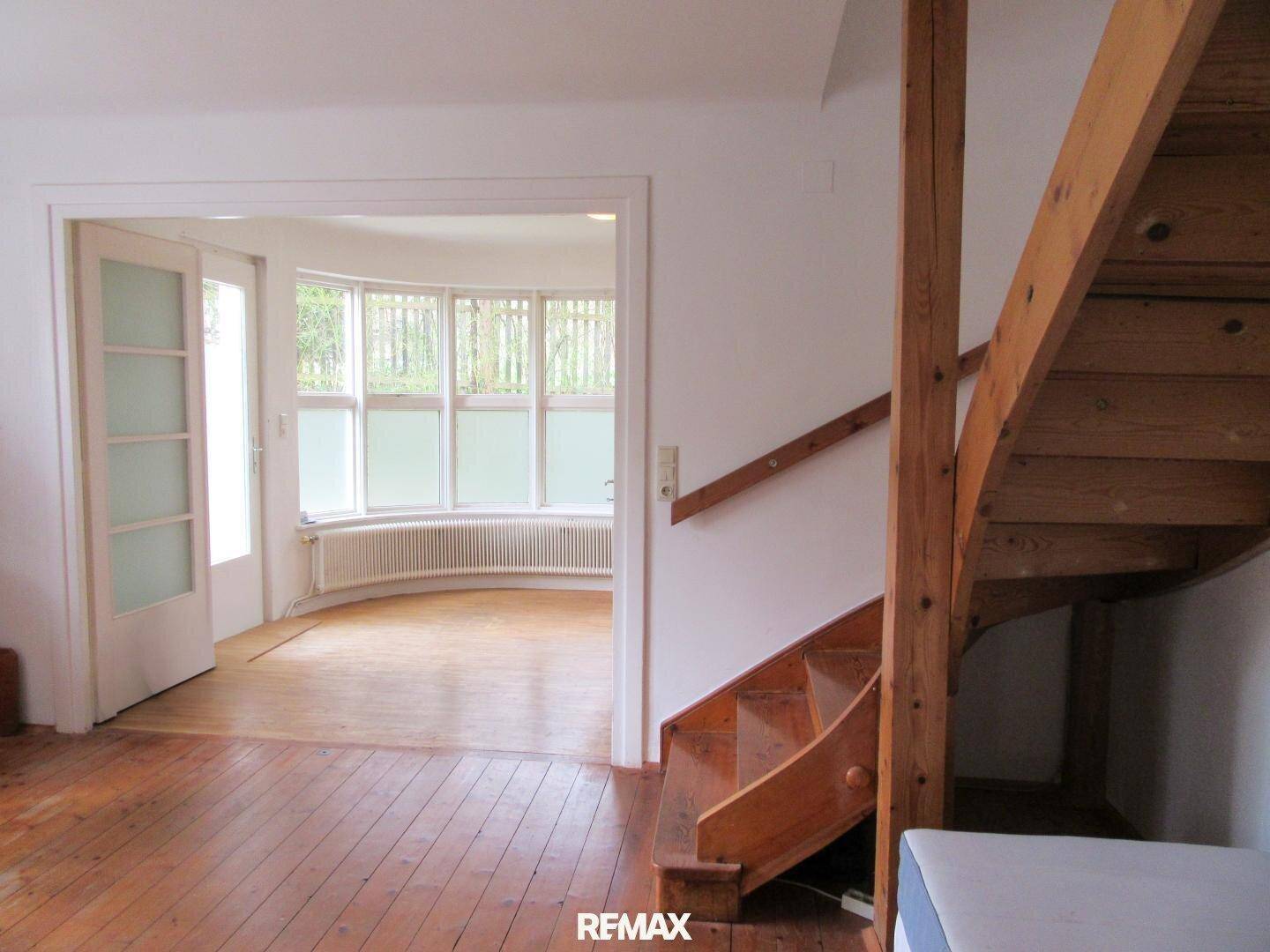 Heller Flurbereich mit Holztreppe und Zugang zu einem Zimmer mit großen Erkerfenstern und Holzboden.