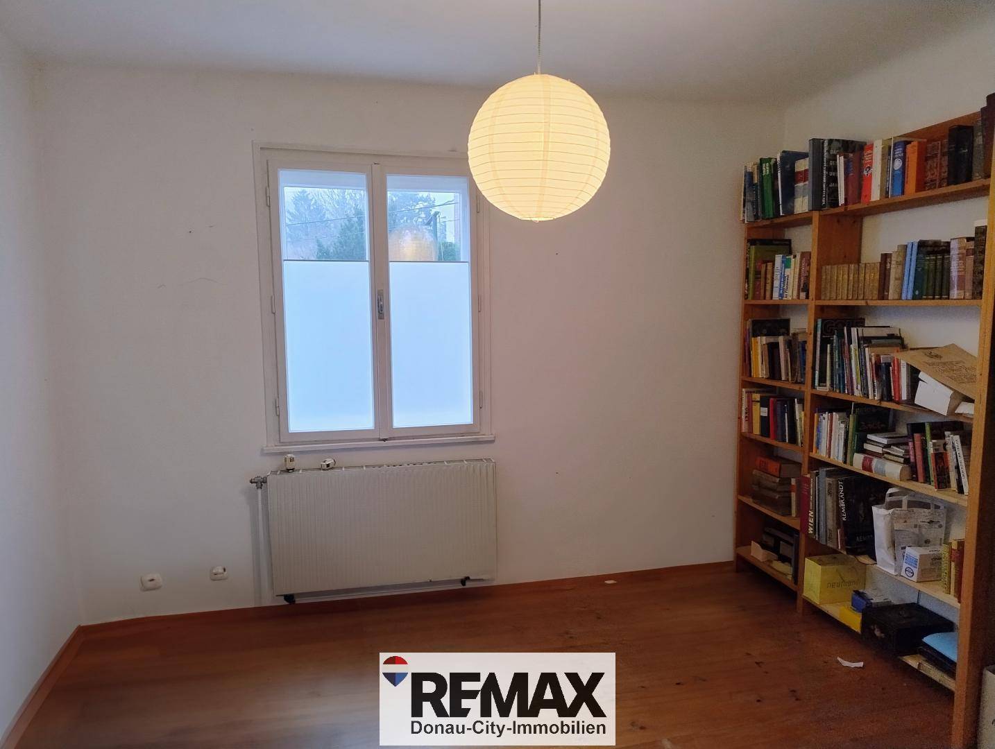 Helles Zimmer mit Holzboden, großem Fenster und einem Bücherregal, ideal als Schlaf- oder Arbeitszimmer.