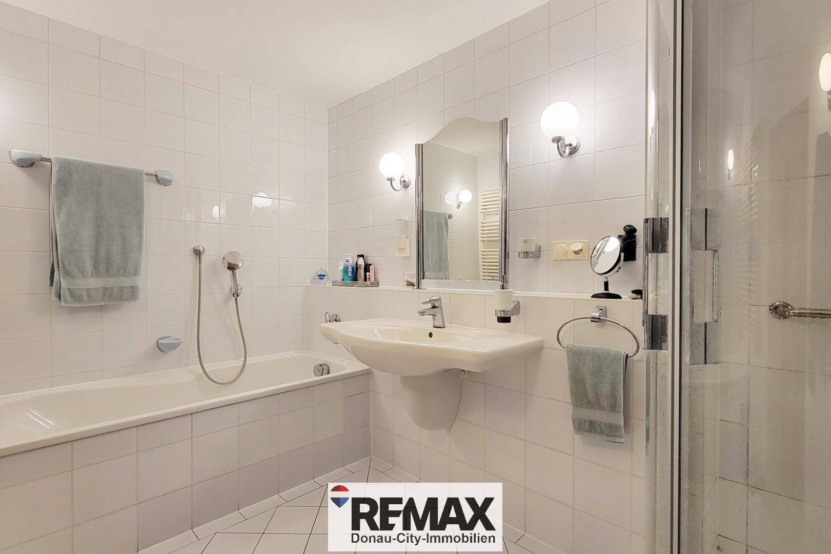 Helles Badezimmer mit weißen Fliesen, Badewanne, Dusche und Einzelwaschbecken.