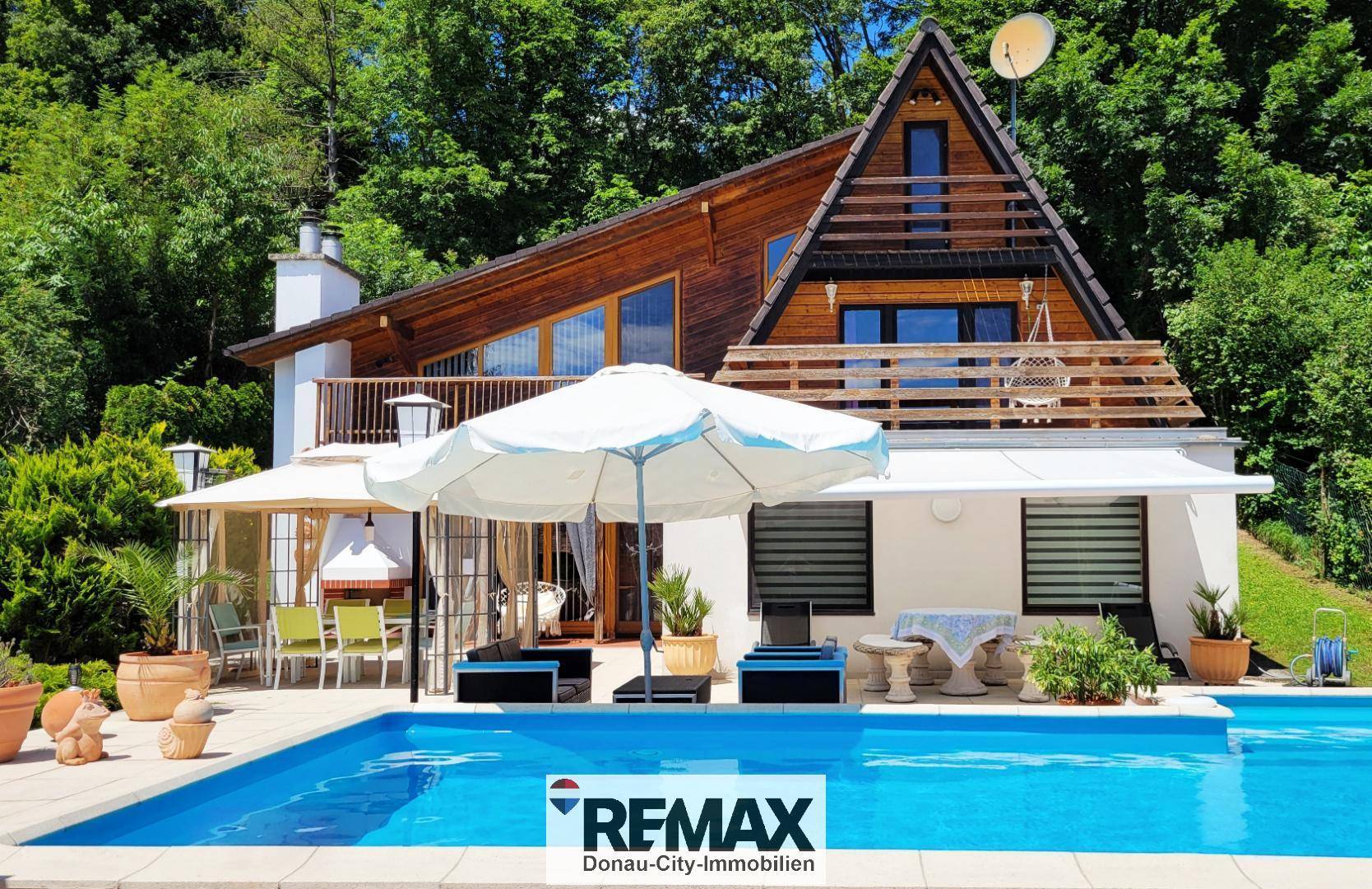 Attraktives Holzhaus mit Poolbereich, Sonnenschirm und Liegestühlen, umgeben von viel Grün.
