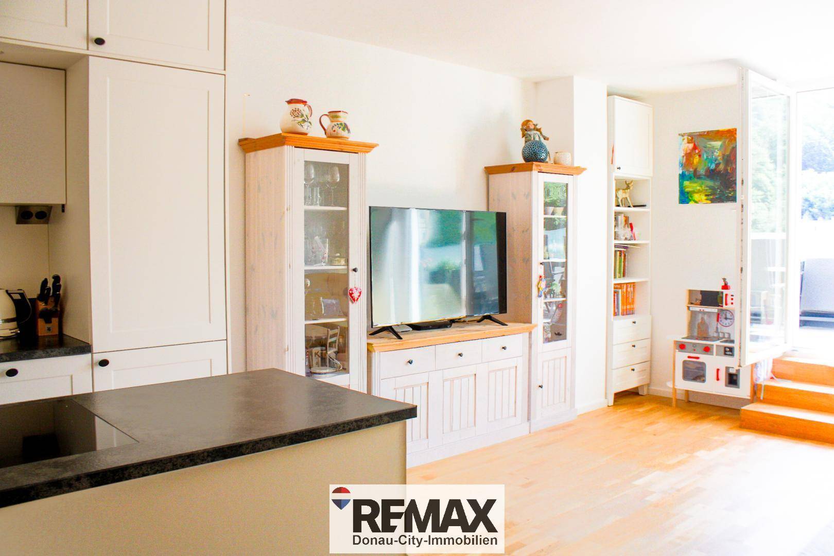 Moderne Küche mit weißen Schränken, Kochinsel und integriertem TV-Bereich, offen zum Wohnraum.