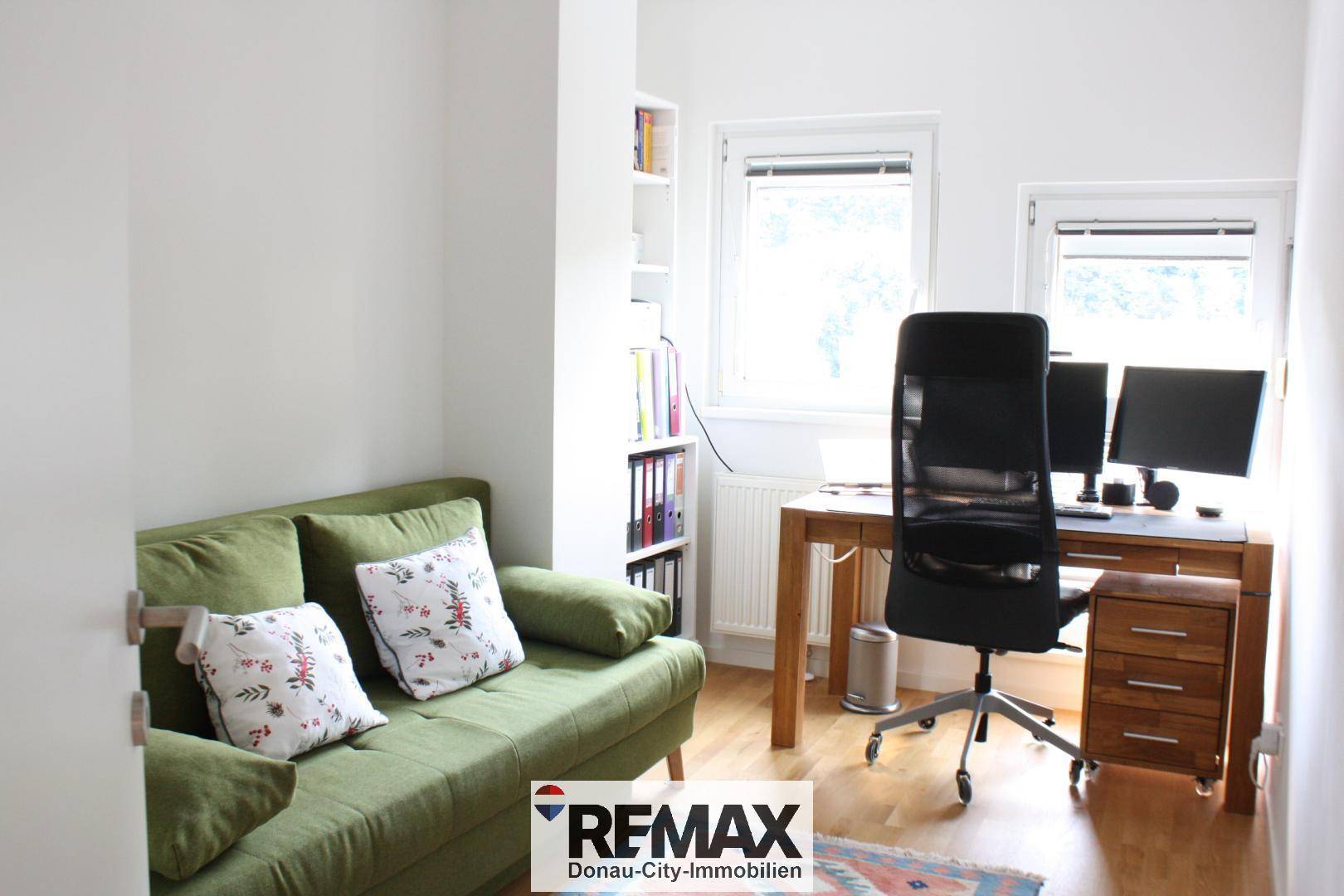 Gemütliches Arbeitszimmer mit grünem Sofa, Holzschreibtisch und Regalen, hell durch zwei Fenster.