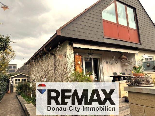 Hausfassade mit Terrasse, Gartenhaus und Dachfenstern unter bewölktem Himmel.