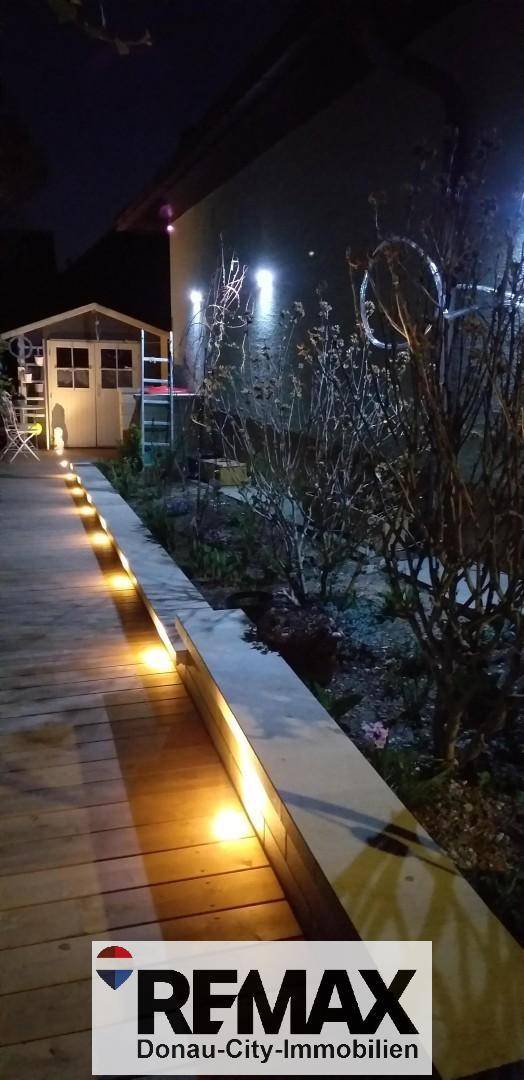Beleuchteter Holzweg entlang der Hauswand mit Gartenhaus im Hintergrund.