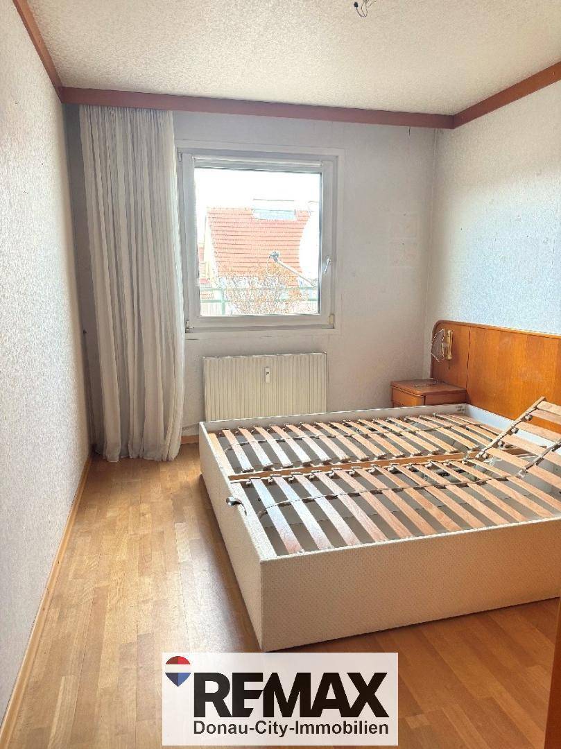 Schlafzimmer mit Holzbettrahmen und Parkettboden, hell durch das Fenster mit Blick nach draußen.