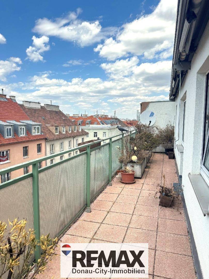 Langer Balkon mit Fliesenboden und Pflanzen, bietet einen weiten Blick über die Dächer der Stadt.