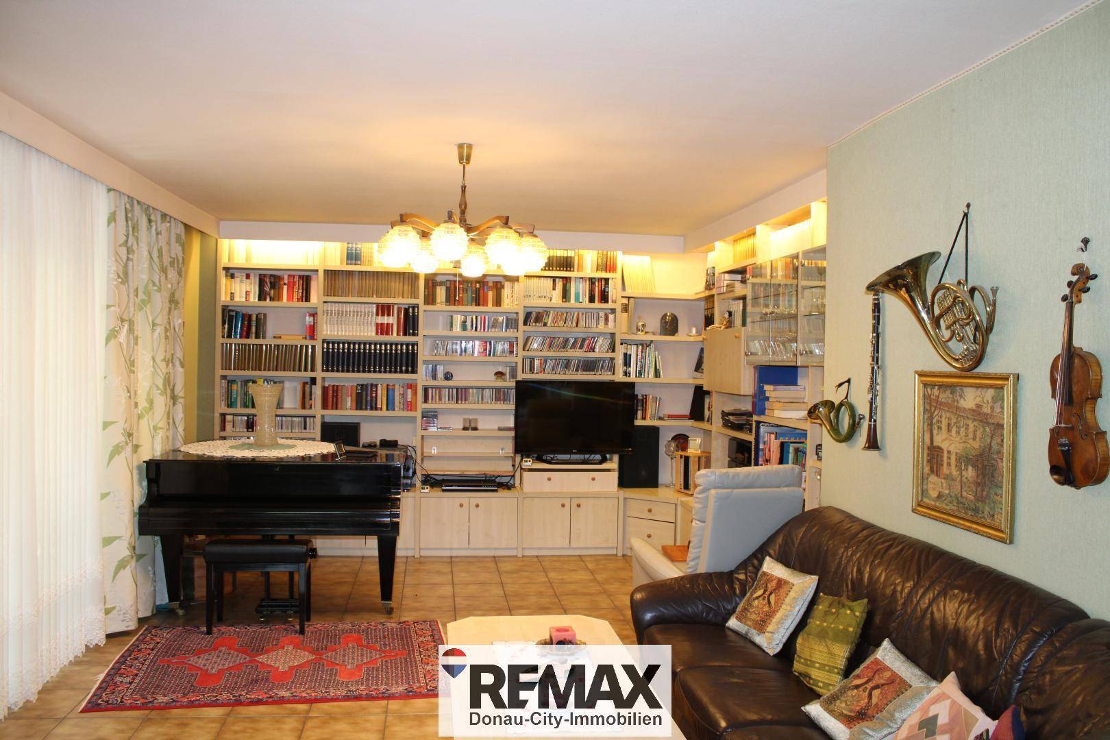 Geräumiges Wohnzimmer mit Ledersofa, Bücherregalen und einem Flügel, beleuchtet von einer Deckenlampe.